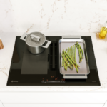 Placa de inducción en funcionamiento, con una olla y una plancha con espárragos, sobre encimera marmolada con utensilios de cocina sobre ella.