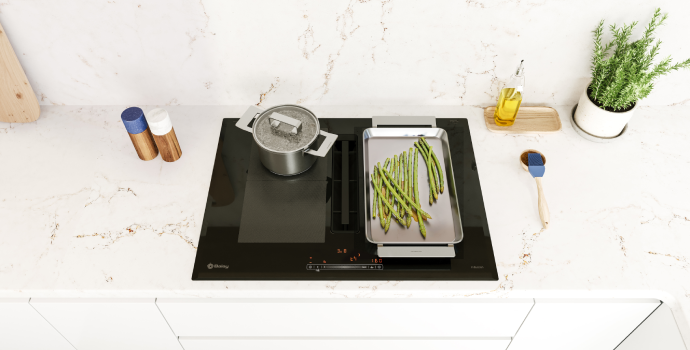 Placa de inducción en funcionamiento, con una olla y una plancha con espárragos, sobre encimera marmolada con utensilios de cocina sobre ella.